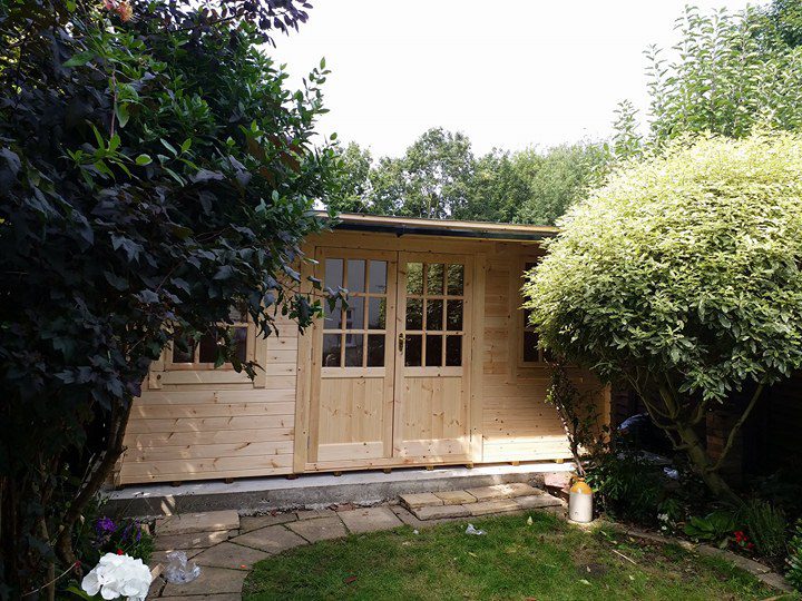 Log cabin with double doors and two windows, in a customer's garden with trees either side.
