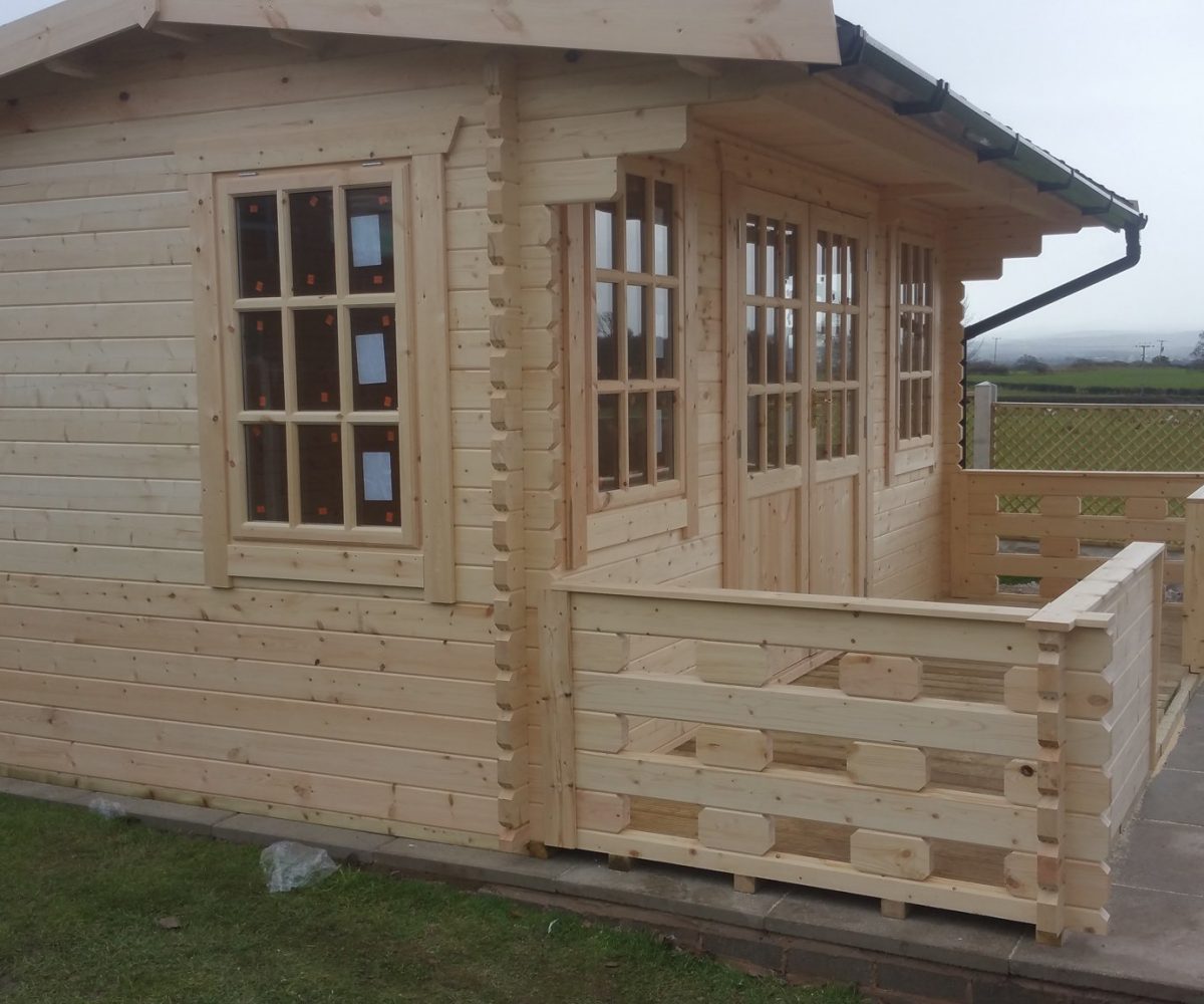 Newly constructed apex roof log cabin with double doors, veranda, and front and side windows, in a garden.