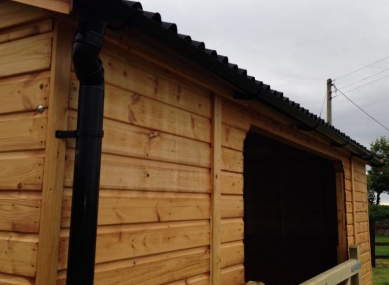 Wooden field shelter with wooden gate, black pipe and felt apex roof, situated in farm yard.
