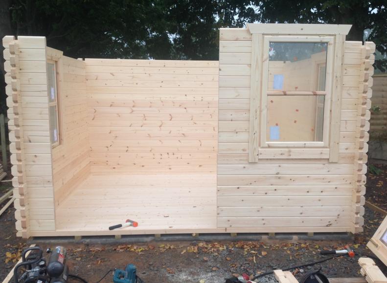 Log cabin mid-construction with walls and window fitted, and a gap for doors, surrounded by tools.