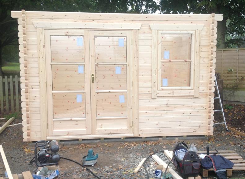 Log cabin mid-construction with walls, fully glazed double doors and window fitted, surrounded by tools.