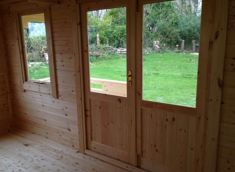 Interior view of log cabin showing half glazed double doors with a window next to them, looking out onto a lawn.